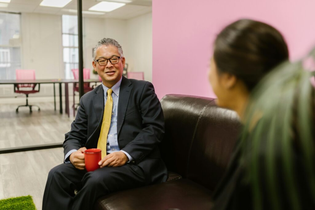 Asian businessman in suit holding red mug during informal office meeting, sitting on a couch.