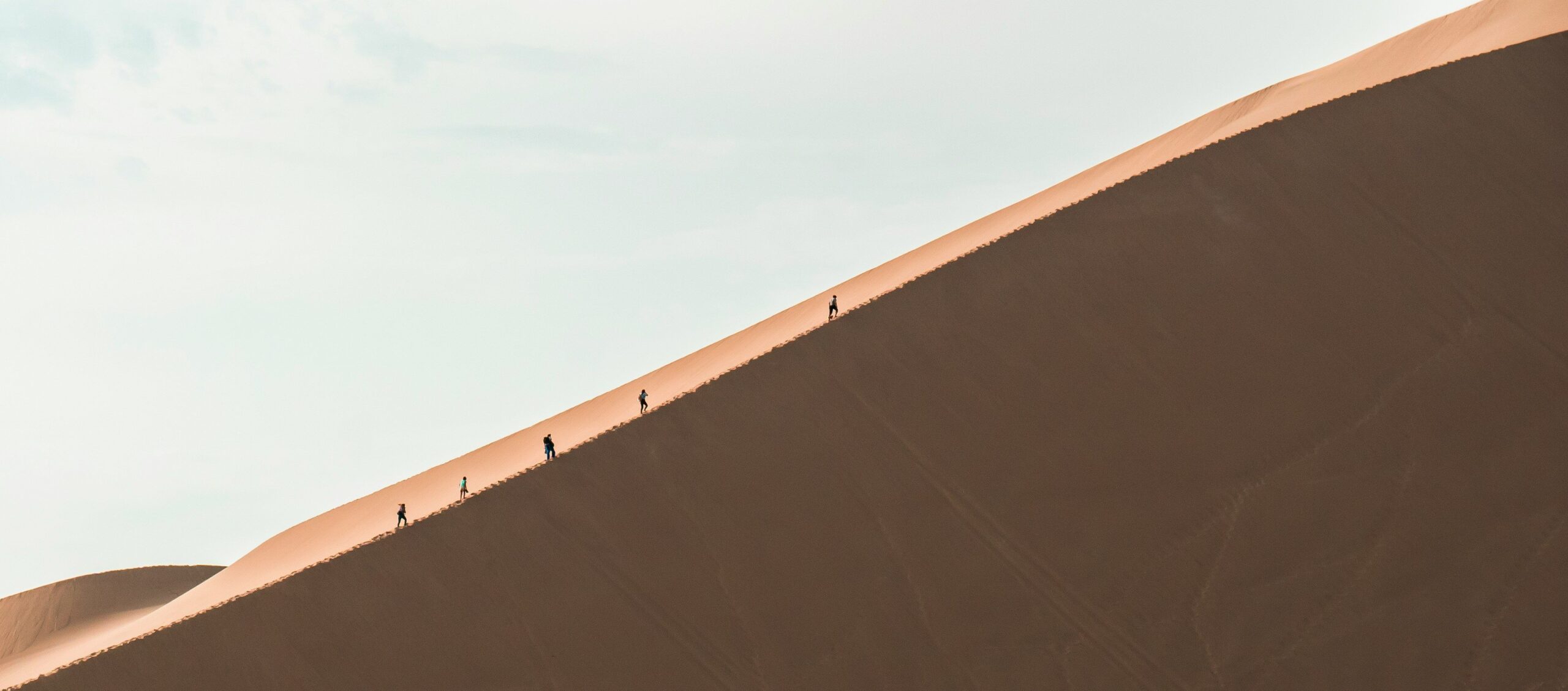 A group of hikers walking up a vast sand dune under a clear sky, showcasing adventure and travel.